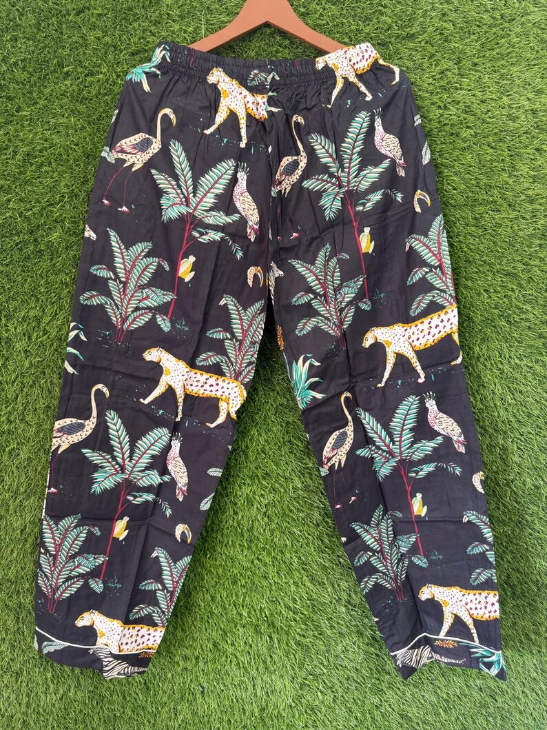 以下が含まれることがあります： ヒョウ、フラミンゴ、熱帯植物の繰り返し模様が施された黒いパジャマパンツ。パンツの裾には白い縁取りがあり、木製のハンガーに吊るされています。生地はコットンに見えます。
