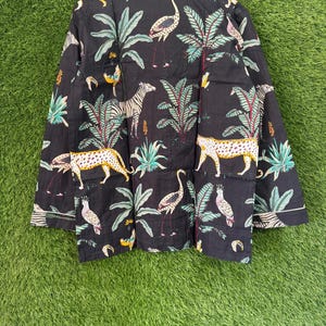 以下が含まれることがあります： 黒色のシャツで、シマウマ、ヒョウ、フラミンゴ、オウムが描かれたジャングル柄です。デザインには緑の葉と白いトリムが含まれています。シャツは木製のハンガーに吊るされています。