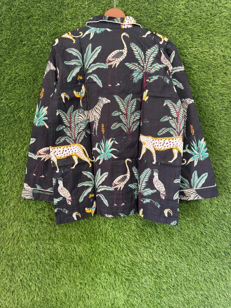 以下が含まれることがあります： 黒色のシャツで、シマウマ、ヒョウ、フラミンゴ、オウムが描かれたジャングル柄です。デザインには緑の葉と白いトリムが含まれています。シャツは木製のハンガーに吊るされています。