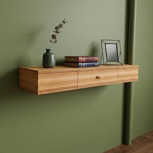May include: A light brown wooden floating shelf with a drawer, holding a small green vase with a plant, two books, and a black framed picture. The shelf is mounted on a green wall.