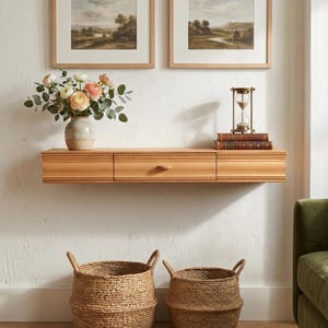May include: A light wood floating shelf with three drawers, a vase of flowers, and two framed landscape paintings. Two woven baskets sit on the floor below. An hourglass and books are on the shelf. A green sofa is on the right.