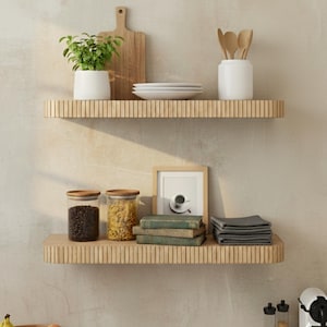 May include: Two wooden shelves with various kitchen items. The top shelf holds a cutting board, white plates, a plant in a white pot, and a white utensil holder with wooden spoons. The bottom shelf has jars, books, and folded gray cloths.