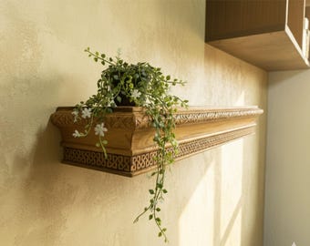 Può includere: Una mensola decorativa in legno con intagli intricati e una pianta in vaso. La mensola è montata su una parete di colore chiaro. La pianta ha foglie verdi e piccoli fiori bianchi, aggiungendo un tocco di natura all'arredamento.