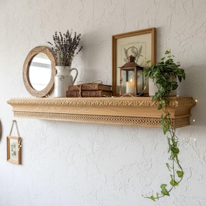 May include: A light-colored wooden shelf with decorative trim, holding a mirror, a vase of lavender, books, a framed botanical print, a lantern with a lit candle, and a potted plant. The shelf is mounted on a white textured wall, creating a rustic, yet elegant display.