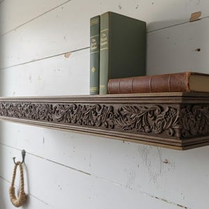 Op de afbeelding: Een donkerbruine houten zwevende plank met gedetailleerde bloem- en bladmotieven uitgesneden langs de voorrand. Twee groene boeken met harde kaft staan rechtop en één bruin in leer gebonden boek ligt plat op de plank.