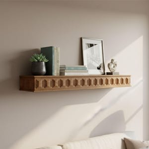 Può includere: Una mensola galleggiante in legno con un design decorativo intagliato. La mensola contiene una cornice, libri, una piccola pianta grassa in vaso e una piccola scultura. La mensola è montata su una parete di colore neutro.