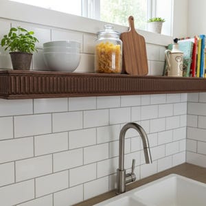 May include: A dark brown, rectangular floating shelf with a grooved design, mounted on a white tiled wall. The shelf holds a potted plant, white bowls, a jar of pasta, a wooden cutting board, and books. A stainless steel faucet is in the foreground.