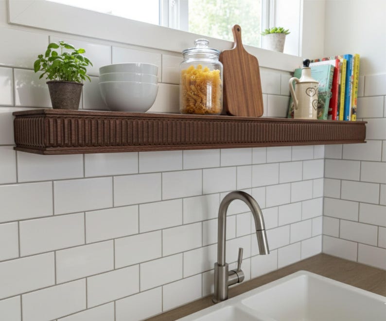 May include: A dark brown, rectangular floating shelf with a grooved design, mounted on a white tiled wall. The shelf holds a potted plant, white bowls, a jar of pasta, a wooden cutting board, and books. A stainless steel faucet is in the foreground.