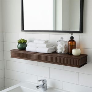 May include: A bathroom shelf with a dark brown finish, holding white towels, a green potted plant, a glass jar, a brown bottle with a black pump, and a white candle. A black-framed mirror hangs above.