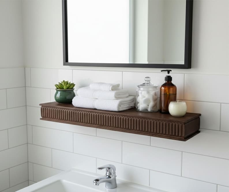 May include: A bathroom shelf with a dark brown finish, holding white towels, a green potted plant, a glass jar, a brown bottle with a black pump, and a white candle. A black-framed mirror hangs above.