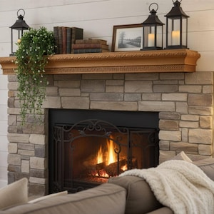 Op de afbeelding: Een gezellige open haard met een stenen gevel en een houten mantel. De mantel toont boeken, ingelijste kunst en twee zwarte lantaarns met brandende kaarsen. Een groene plant hangt naar beneden en er brandt een vuur in de haard.