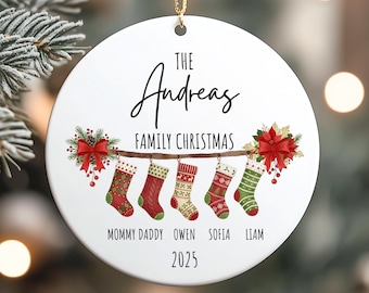 May include: A white Christmas ornament with a gold string, featuring the text "The Andreas Family Christmas" and the year 2025. It displays five hanging Christmas stockings in red and green, personalized with names.