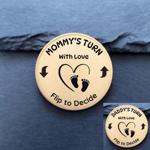 May include: A gold-coloured coin with the text "MOMMY'S TURN" and "DADDY'S TURN" in a circular design. The coin features a heart with baby footprints and the phrase "Flip to Decide".