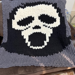 Puede incluir: Una manta de punto grueso hecha a mano con un diseño de calavera. La calavera es blanca sobre un fondo negro, enmarcada por un borde gris. La manta está hecha de hilo grueso y suave, ideal para la decoración del hogar.