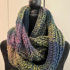 Puede incluir: Bufanda infinita tejida a mano en tonos azules, verdes, amarillos y rosas. La bufanda de punto grueso se exhibe en una cabeza de maniquí negra. La bufanda es un accesorio acogedor para el clima frío.