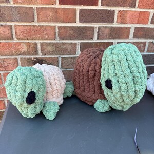 May include: Three handmade crochet turtle plushies in shades of green, brown, and beige. Each turtle has a round body, small legs, and a black eye. The turtles are displayed on a grey surface.