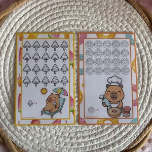 Op de afbeelding: Twee rechthoekige papieren spaartrackers met capybara-illustraties. De linkerkaart heeft watermeloenpictogrammen en een capybara op een ligstoel. De rechterkaart toont donutpictogrammen en een capybara met een koksmuts en spuitzak.