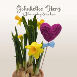 Könnte beinhalten: Ein Topfarrangement aus gelben Narzissen mit grünen Blättern und einem lila gehäkelten Herz an einem Holzstab. Das Herz ist mit einer blauen Schleife verziert. Der Text im Bild lautet "Gehäkeltes Herz Blumenpotfstecken".