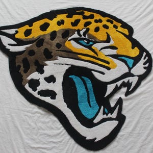 May include: A rug shaped like a jaguar's head, featuring a black outline, yellow and brown spots, and a blue tongue. The rug is made of a soft, textured material and is designed to resemble the animal's face.