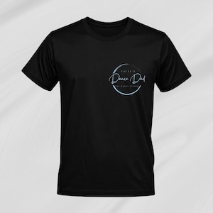 Op de afbeelding: Zwart T-shirt met ronde hals en een lichtblauw cirkelvormig ontwerp op de linkerborst. Het ontwerp bevat de tekst "Lacey's Dance Dad The Dance Studio" in een handschriftachtig lettertype. Het shirt is gemaakt van een gladde, matte stof.