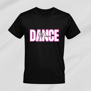 May include: Black t-shirt featuring the text "DANCE Mom" in a white and pink block letter design. The word "DANCE" is in a bold, sans-serif font, while "Mom" is in a cursive script. The shirt has a crew neck.