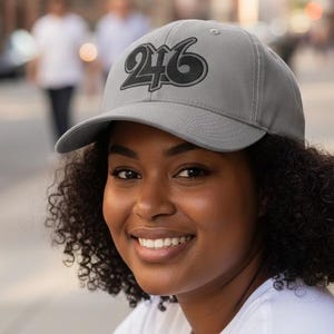 May include: A gray baseball cap with the number "246" in black script. The cap is worn by a person wearing a white t-shirt and blue jeans. The background is a city street with blurred figures.