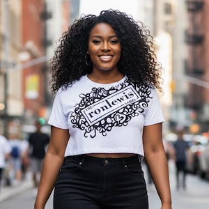 May include: A woman wearing a white crop top with the text "ronlewis" in a black decorative design, paired with black denim shorts. She is wearing white sandals and walking on a city street.