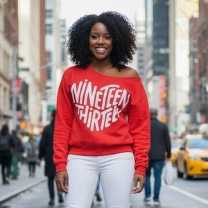 May include: A woman wearing a red off-the-shoulder sweatshirt with the text "NINETEEN THIRTEE" in white, white skinny jeans, and red heels. The photo was taken on a city street with people and yellow taxis in the background.