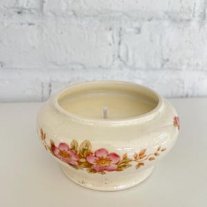 May include: Cream-colored candle in a decorative ceramic bowl. The bowl features a floral pattern with pink flowers and green leaves. The candle has a single wick and is set against a white brick background.