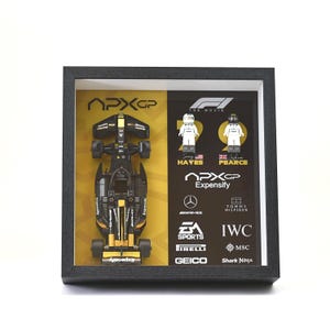 May include: Framed APXGP race car model in black and yellow, with two Lego figures and sponsor logos. The frame is black, and the background is divided into yellow and white sections.
