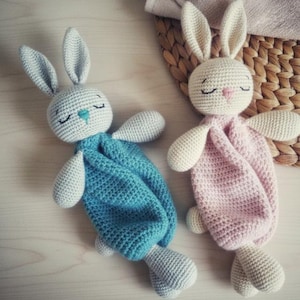 Puede incluir: Dos juguetes de conejito de ganchillo hechos a mano. Uno es azul, el otro rosa. Ambos tienen orejas largas, ojos cerrados y narices pequeñas. Los conejitos están sobre una superficie de color claro.