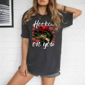 Może przedstawiać: Ciemnoszara koszulka z grafiką okonia w okularach przeciwsłonecznych i czerwonej czapce, otoczona czerwonymi odciskami warg. Tekst "Hooked on you" znajduje się nad i pod rybą. Koszulka ma swobodny styl.