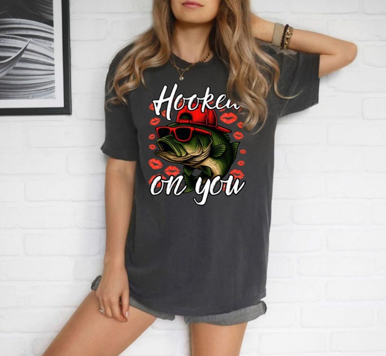 Może przedstawiać: Ciemnoszara koszulka z grafiką okonia w okularach przeciwsłonecznych i czerwonej czapce, otoczona czerwonymi odciskami warg. Tekst "Hooked on you" znajduje się nad i pod rybą. Koszulka ma swobodny styl.