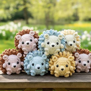 Puede incluir: Colección de peluches de león hechos a mano con ganchillo en varios colores, incluyendo rosa, azul, amarillo y marrón. Cada león tiene un cuerpo redondo, una melena esponjosa y pequeños ojos negros. Los juguetes están colocados sobre una superficie de madera.