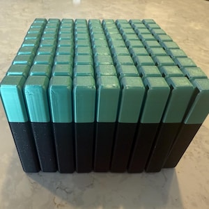May include: A rectangular block composed of numerous teal and black rectangular prisms. The teal sections are on top, and the black sections are on the bottom. The prisms are arranged in a grid-like pattern, creating a textured surface.