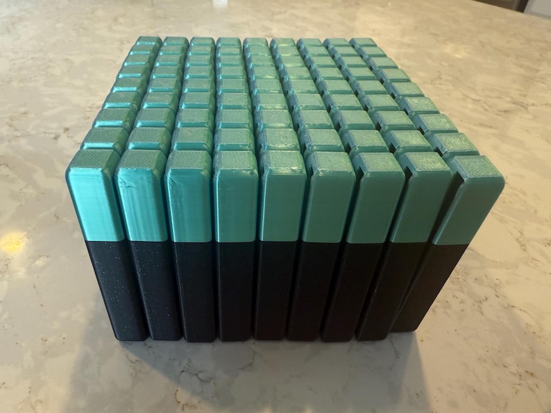 May include: A rectangular block composed of numerous teal and black rectangular prisms. The teal sections are on top, and the black sections are on the bottom. The prisms are arranged in a grid-like pattern, creating a textured surface.