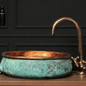 May include: A copper and verdigris patina vessel sink with a bronze faucet. The sink has a rounded shape with a copper rim. The faucet has a curved spout and cross handles. The sink and faucet are on a wooden surface.