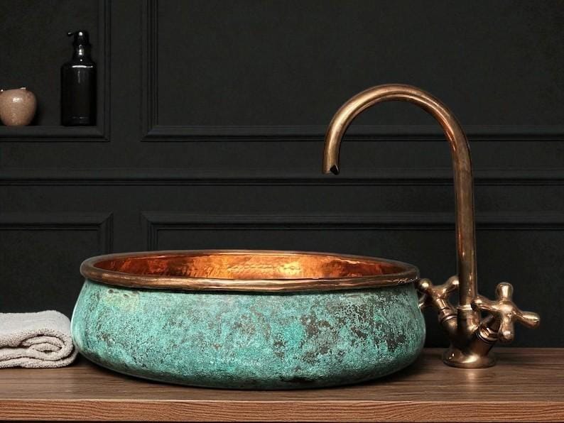May include: A copper and verdigris patina vessel sink with a bronze faucet. The sink has a rounded shape with a copper rim. The faucet has a curved spout and cross handles. The sink and faucet are on a wooden surface.
