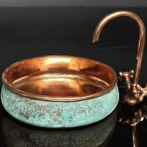May include: A copper sink with a verdigris exterior and a polished interior. A matching copper faucet with cross handles is positioned to the right. The sink has a round shape and sits on a reflective surface.