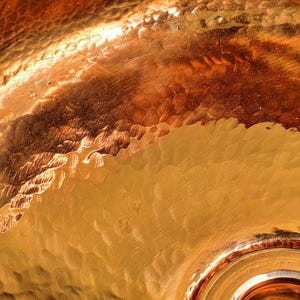 May include: Close-up of a hammered copper surface with a warm, golden-brown hue. The texture shows a pattern of irregular, small indentations and a smooth, reflective finish. The light reflects off the surface, creating highlights and shadows.