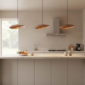 Puede incluir: Tres l&aacute;mparas colgantes de color cobre sobre una isla de cocina. Las luces tienen forma de disco y est&aacute;n suspendidas por finos cables negros. La cocina presenta una paleta de colores neutros con una ventana y electrodom&eacute;sticos de acero inoxidable.