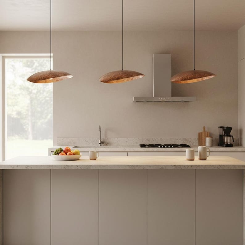 Puede incluir: Tres l&aacute;mparas colgantes de color cobre sobre una isla de cocina. Las luces tienen forma de disco y est&aacute;n suspendidas por finos cables negros. La cocina presenta una paleta de colores neutros con una ventana y electrodom&eacute;sticos de acero inoxidable.