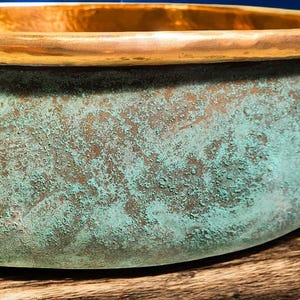 May include: Close-up of a copper bowl with a weathered, textured surface. The bowl's exterior features a turquoise patina over a bronze base, with a polished gold rim. The bowl sits on a wooden surface.
