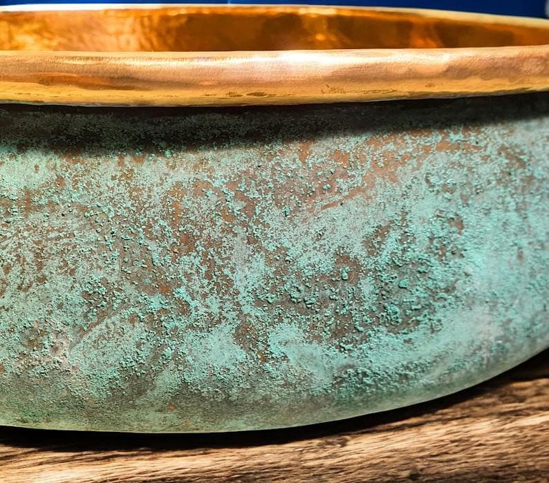 May include: Close-up of a copper bowl with a weathered, textured surface. The bowl's exterior features a turquoise patina over a bronze base, with a polished gold rim. The bowl sits on a wooden surface.