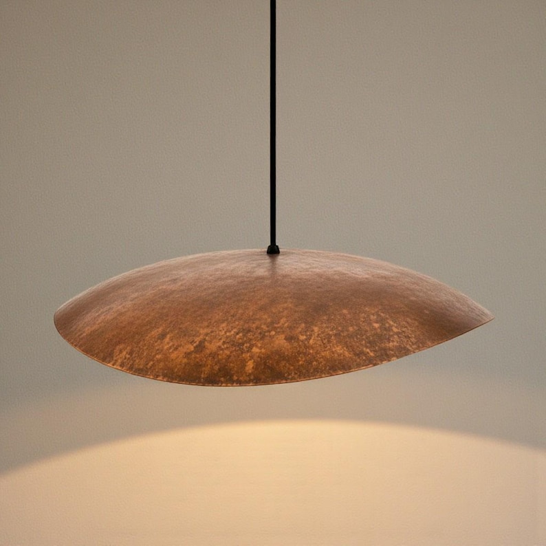 Puede incluir: Una l&aacute;mpara colgante de metal martillado de color cobre. La l&aacute;mpara tiene una pantalla ancha y poco profunda, en forma de platillo, suspendida de un cable negro. La pantalla tiene una superficie texturizada y un tono c&aacute;lido y terroso.