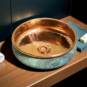 May include: A hammered copper sink with a verdigris exterior and a polished copper interior. The round vessel sink has a decorative drain and sits on a wooden surface. A rolled white towel and a bar of soap are visible.