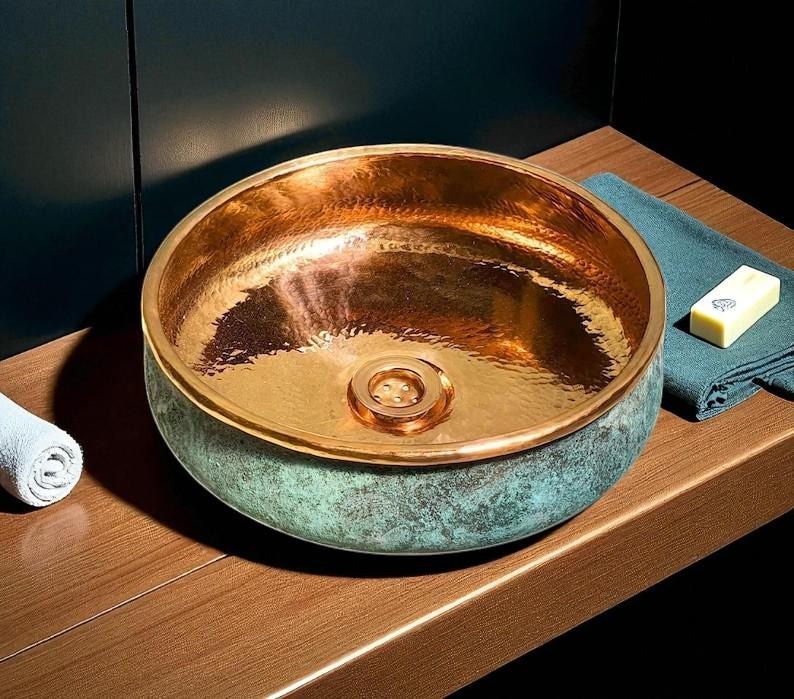 May include: A hammered copper sink with a verdigris exterior and a polished copper interior. The round vessel sink has a decorative drain and sits on a wooden surface. A rolled white towel and a bar of soap are visible.