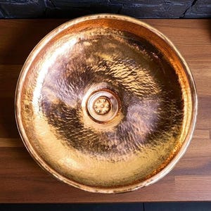 May include: A hammered copper sink with a round basin. The sink has a textured surface and a drain in the center. The copper has a warm, golden hue, reflecting light. The sink is designed for bathroom or kitchen use.