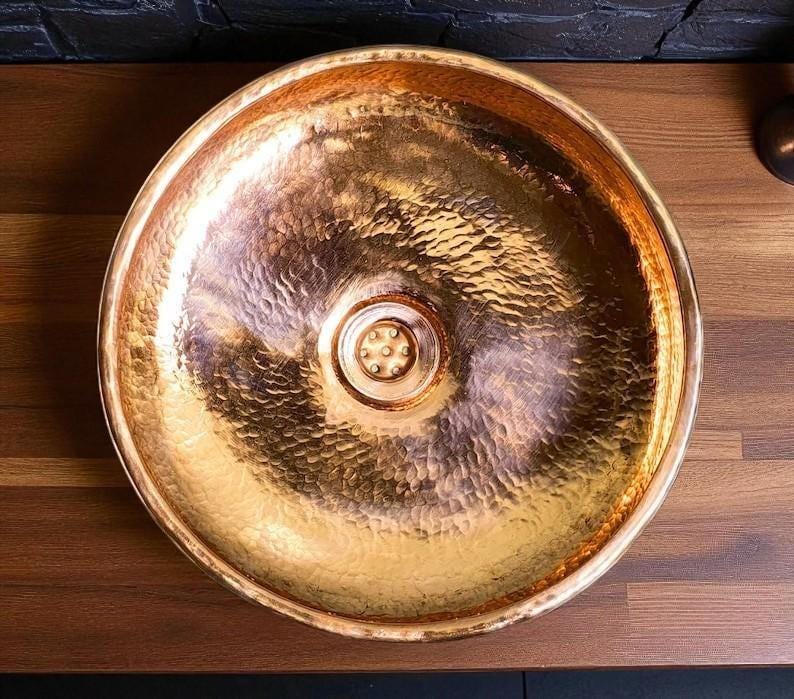 May include: A hammered copper sink with a round basin. The sink has a textured surface and a drain in the center. The copper has a warm, golden hue, reflecting light. The sink is designed for bathroom or kitchen use.