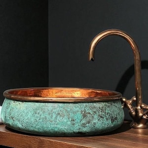 May include: A copper and verdigris patina basin with a bronze faucet. The basin has a rounded shape with a copper rim. The faucet has a curved spout and cross-shaped handles. A rolled white towel sits to the left.